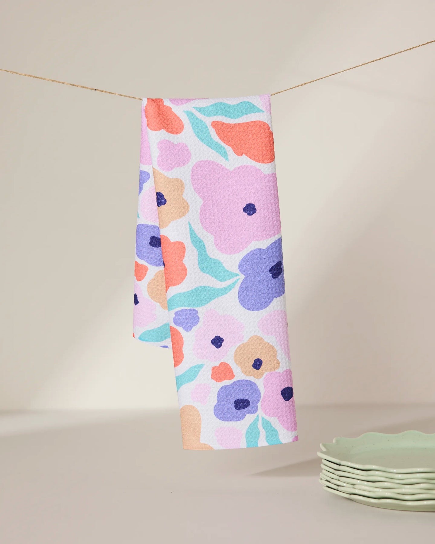The GEOMETRY Blossoming Love tea towel, made from recycled materials and featuring a colorful floral pattern, hangs on a string against a neutral background with light green scalloped plates stacked nearby.