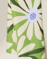 The Bliss and Bloom kitchen towel by GEOMETRY features a cream-colored, textured recycled fabric with a bold floral design of green leaves, petals, and a lavender center accented by dark purple dots.