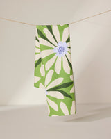 The GEOMETRY Bliss and Bloom tea towel, featuring a bold green and white daisy floral pattern, hangs neatly over a clothesline against a softly lit plain background.