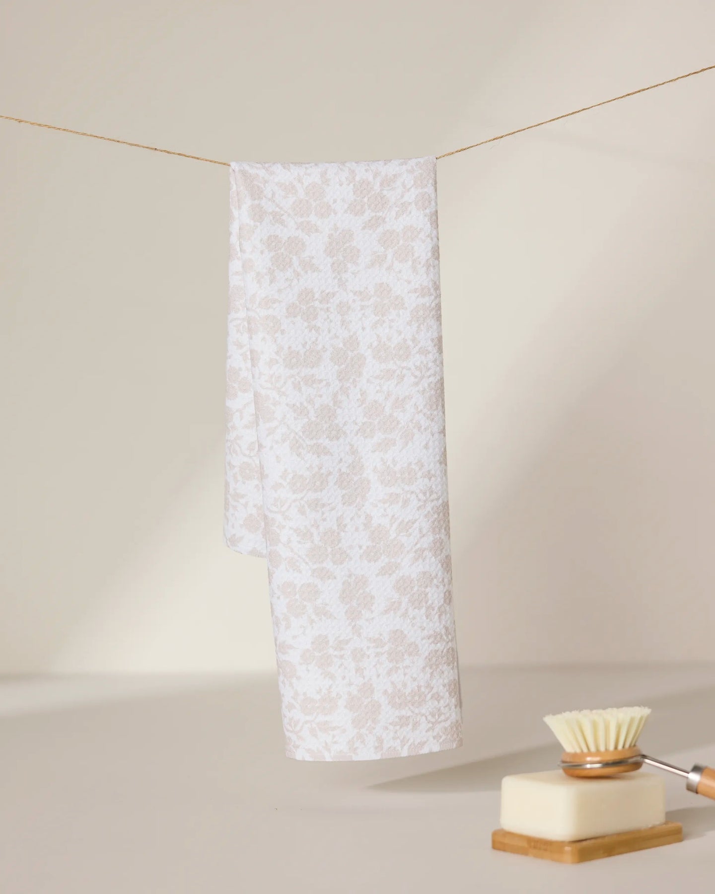 A light beige and white floral-patterned GEOMETRY tea towel, Bettys Buds, hangs on a string against a neutral background. In the foreground, an ultra-absorbent dish brush and a bar of soap rest on a dish.