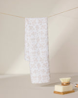 A light beige and white floral-patterned GEOMETRY tea towel, Bettys Buds, hangs on a string against a neutral background. In the foreground, an ultra-absorbent dish brush and a bar of soap rest on a dish.