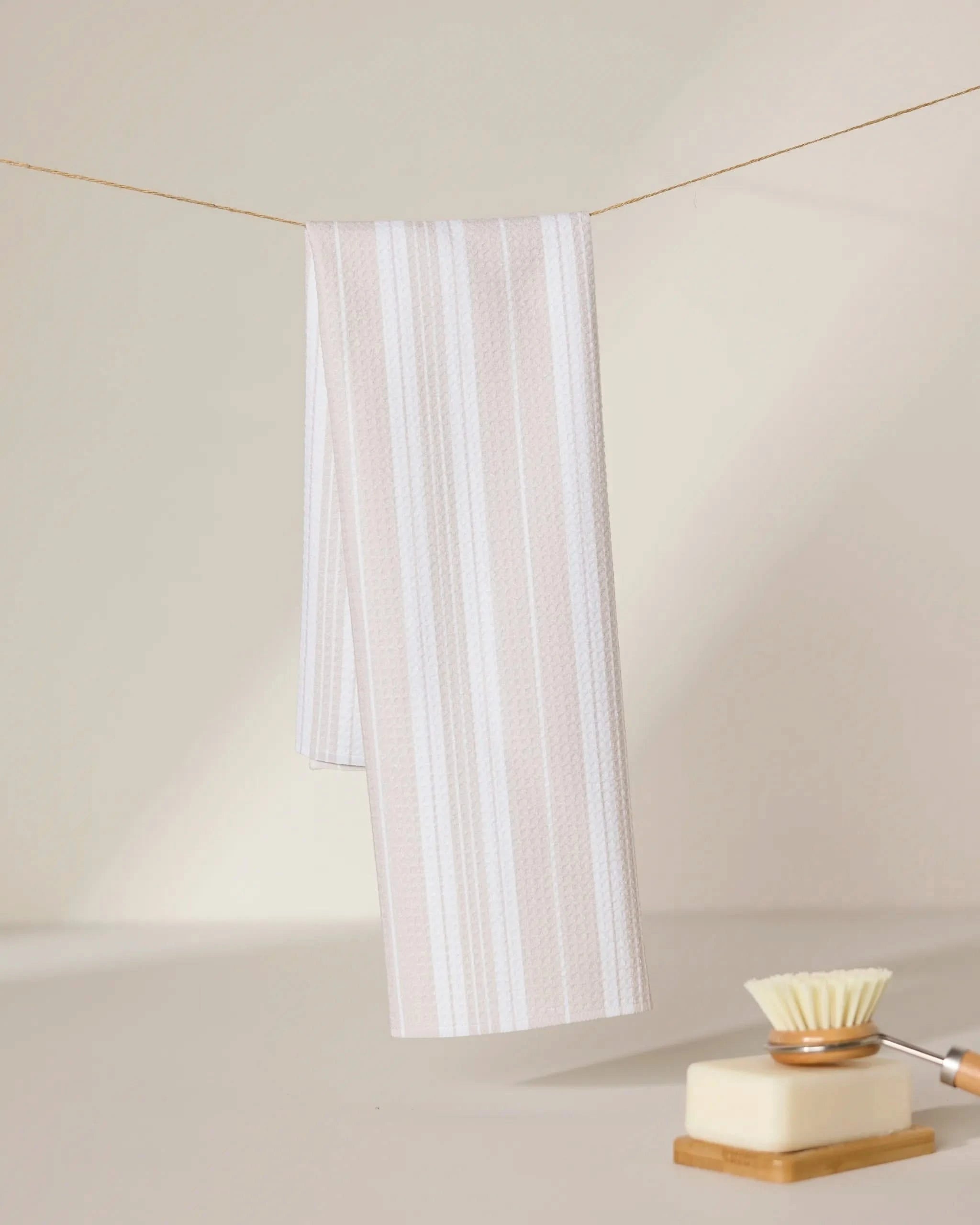 The GEOMETRY Betty Beach striped kitchen towel hangs on a clothesline, with a soap bar and bristled cleaning brush nearby. A softly lit, neutral background highlights the towels serene, beige-and-white design.