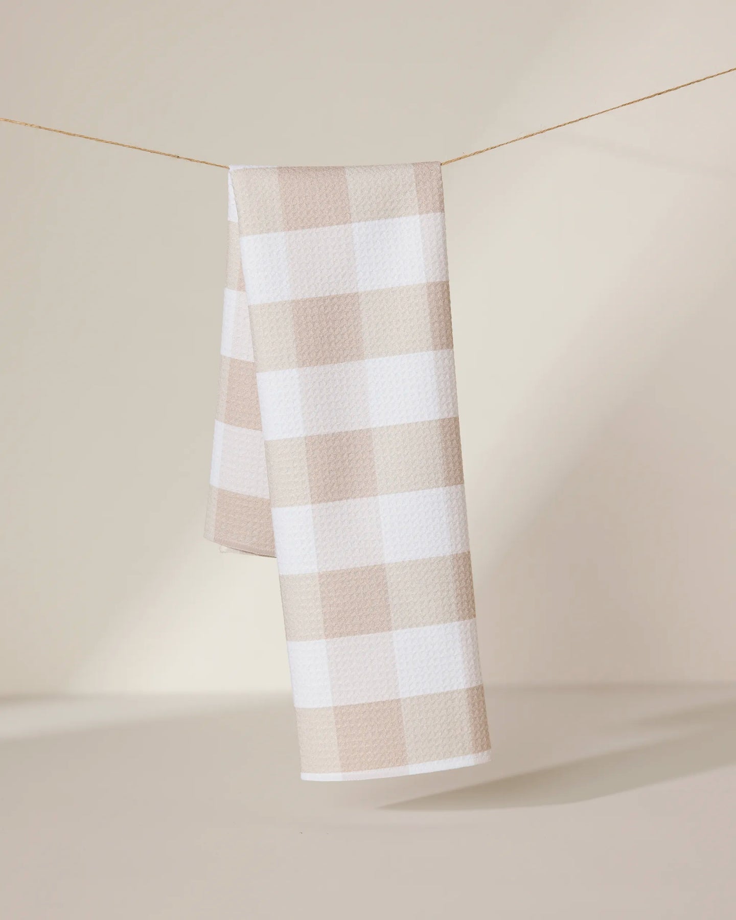 The GEOMETRY Betty Bakes eco-friendly kitchen towel in beige and white checkered design hangs neatly over a thin clothesline, set against a softly lit neutral background.
