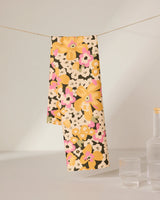 The GEOMETRY Beloved Bunch tea towel, featuring a vibrant floral design in yellow, pink, and white, hangs over a string near two clear glasses and part of a bottle on a plain surface.