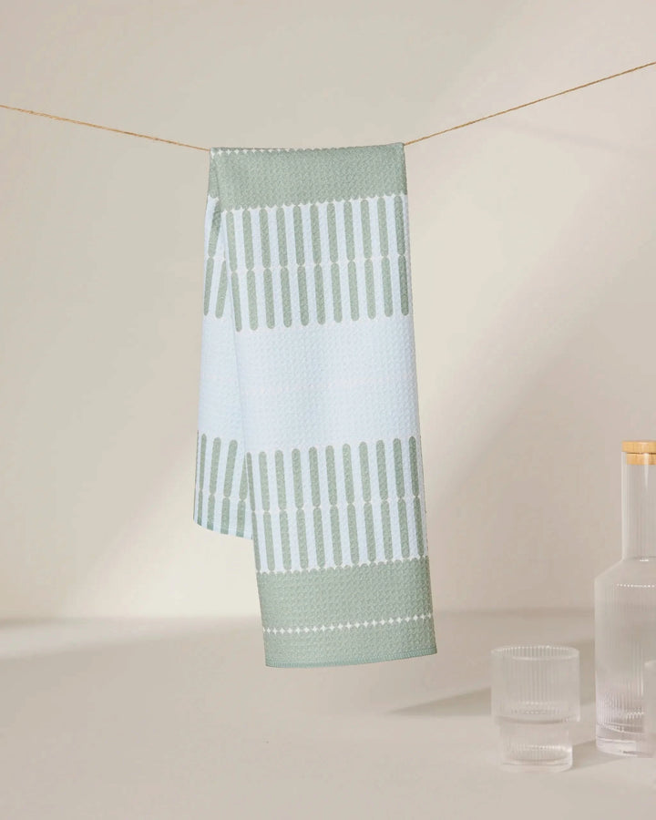 The Bâton Vert tea towel by GEOMETRY, featuring light blue and green patterns and made from recycled materials, hangs on a string with a clear glass bottle and two matching glasses nearby against a neutral background.