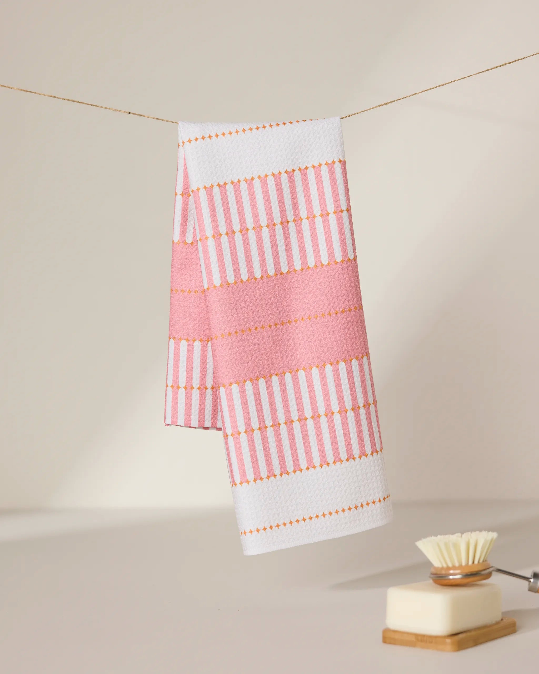 The Bâton Rose tea towel by GEOMETRY, featuring pink and white stripes with orange accents, hangs on a thin line. In the softly lit background, a dish brush with white bristles and a bar of soap rest on the surface.