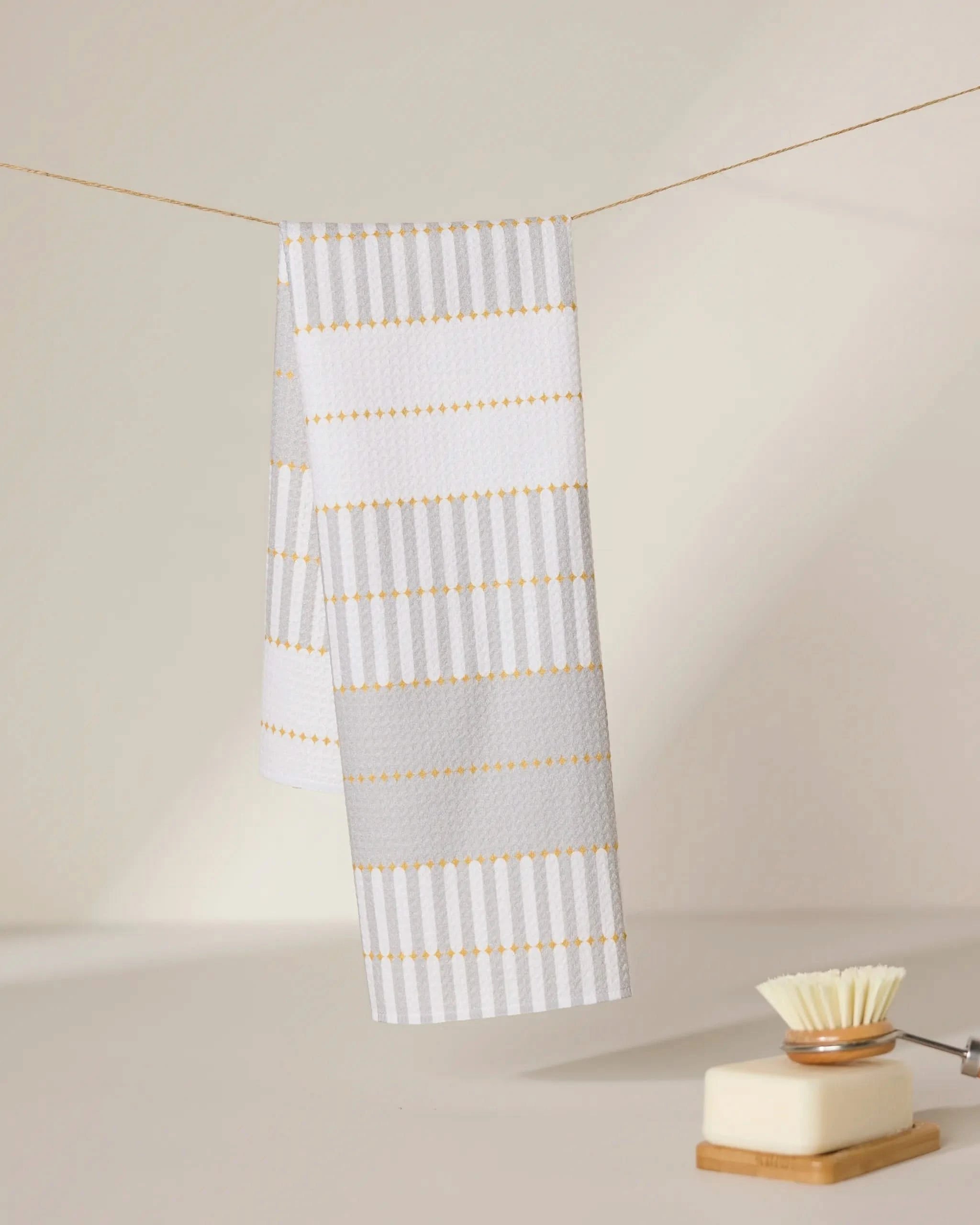 A GEOMETRY Bâton Dor recycled kitchen towel with white and light gray stripes and yellow accents hangs on a line, while a dish brush (white bristle head) and square soap dish sit on a light surface in the foreground.