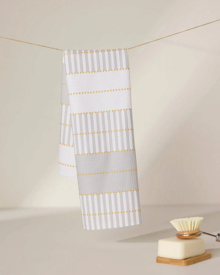 A GEOMETRY Bâton Dor recycled kitchen towel with white and light gray stripes and yellow accents hangs on a line, while a dish brush (white bristle head) and square soap dish sit on a light surface in the foreground.