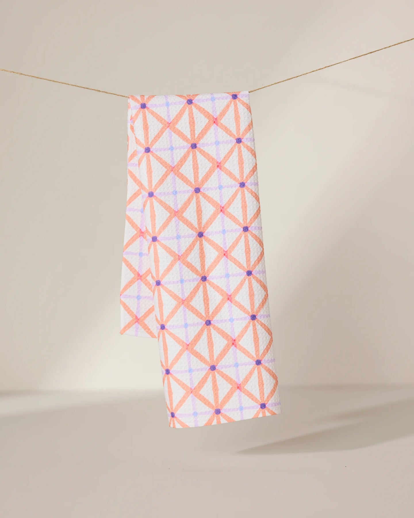 The GEOMETRY Anew tea towel features a quick-drying design with a pink and orange geometric crisscross pattern and small purple dots, shown hanging by a string against a neutral background.