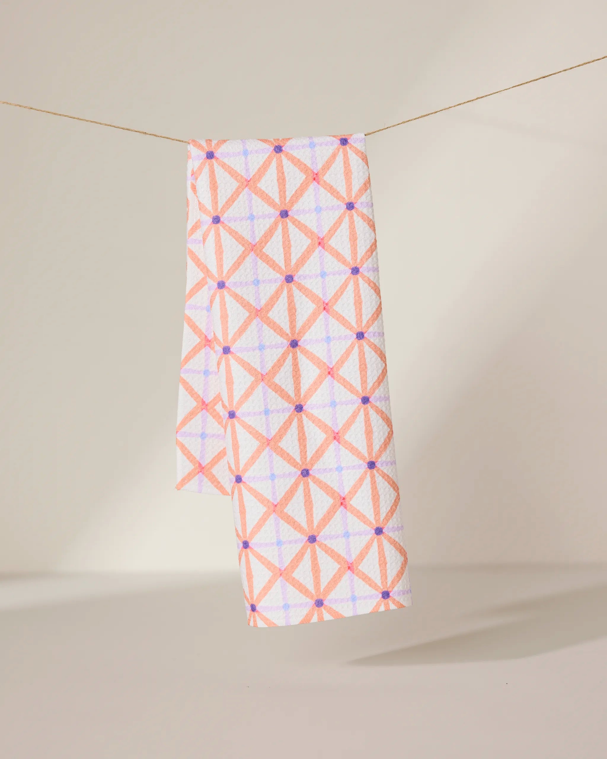 The GEOMETRY Anew tea towel features a quick-drying design with a pink and orange geometric crisscross pattern and small purple dots, shown hanging by a string against a neutral background.