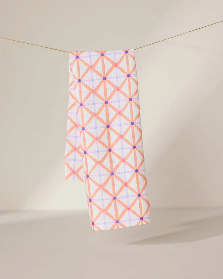 The GEOMETRY Anew tea towel features a quick-drying design with a pink and orange geometric crisscross pattern and small purple dots, shown hanging by a string against a neutral background.