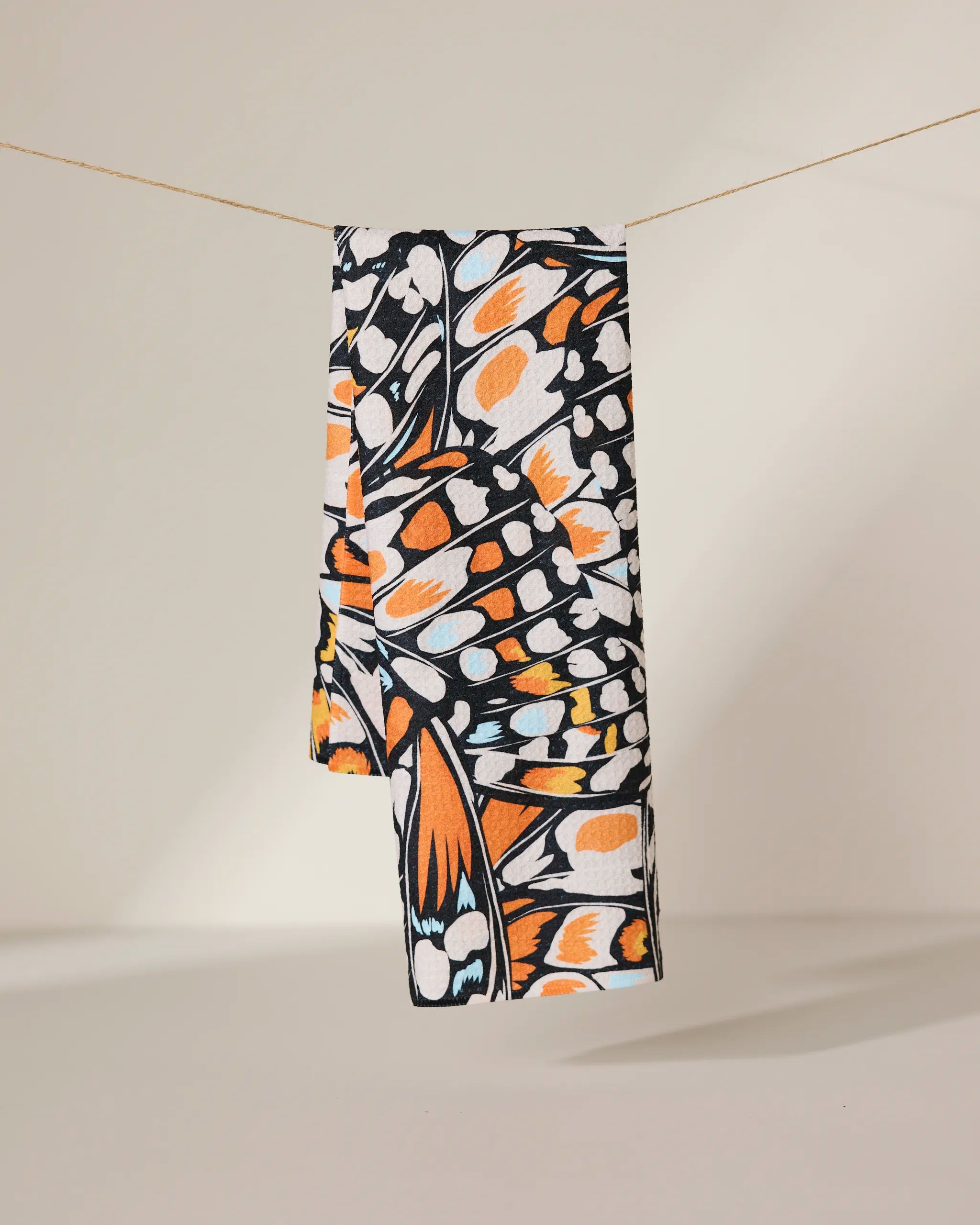 The GEOMETRY Wings in the Wind towel, featuring a vibrant orange, black, and white butterfly wing pattern, hangs neatly on a clothesline against a plain light background.