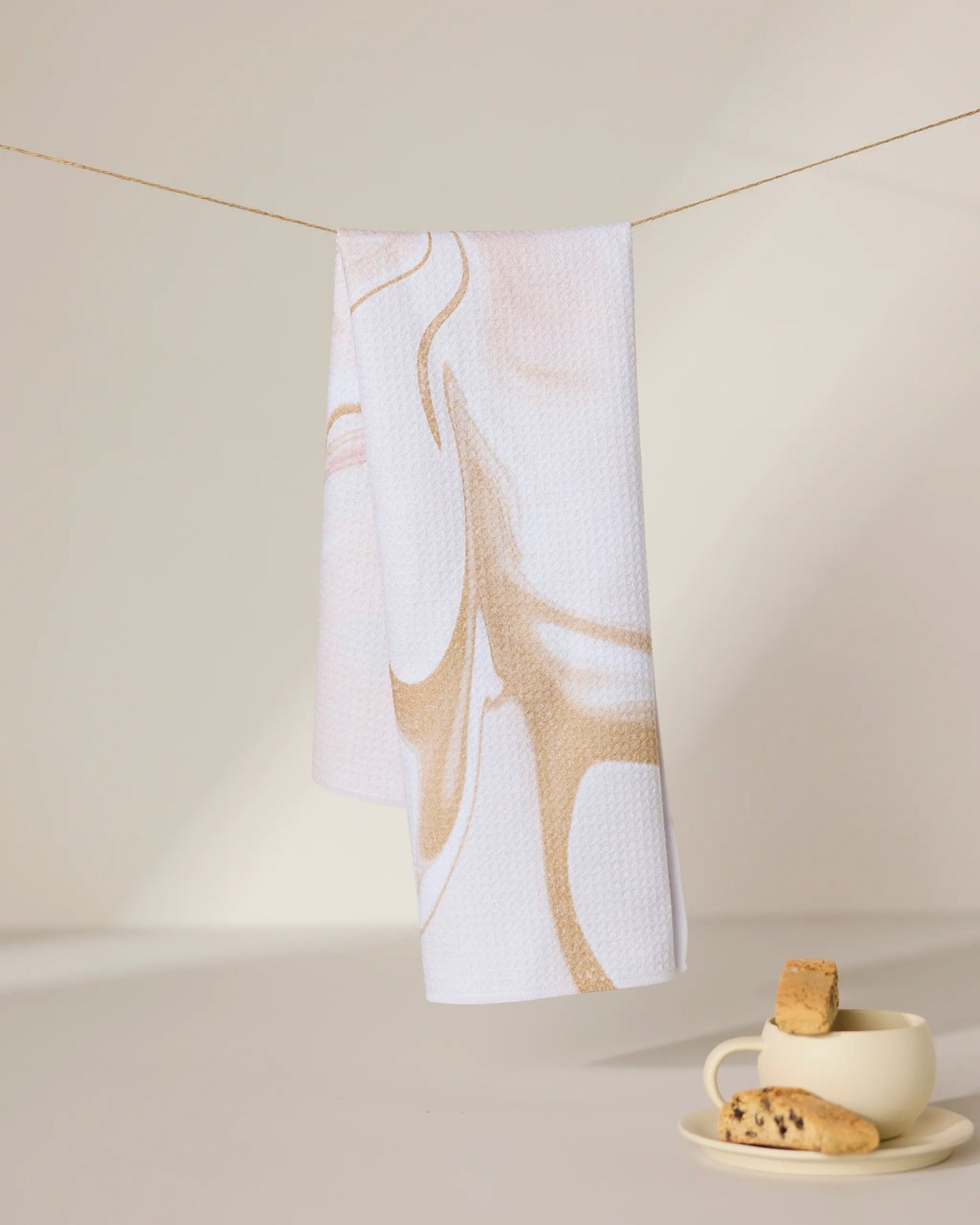 The Vanilla tea towel by GEOMETRY, featuring beige abstract swirls on white and made from recycled materials, hangs above a cream cup and saucer with biscotti on a sunlit surface with soft shadows.