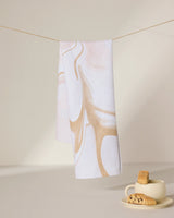 The Vanilla tea towel by GEOMETRY, featuring beige abstract swirls on white and made from recycled materials, hangs above a cream cup and saucer with biscotti on a sunlit surface with soft shadows.