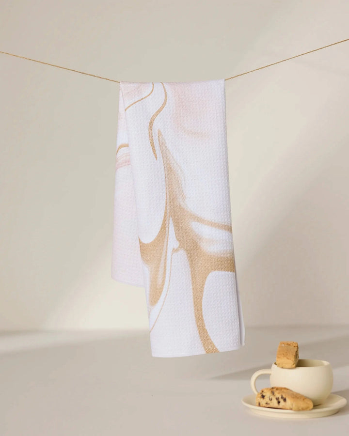 The Vanilla tea towel by GEOMETRY, featuring beige abstract swirls on white and made from recycled materials, hangs above a cream cup and saucer with biscotti on a sunlit surface with soft shadows.