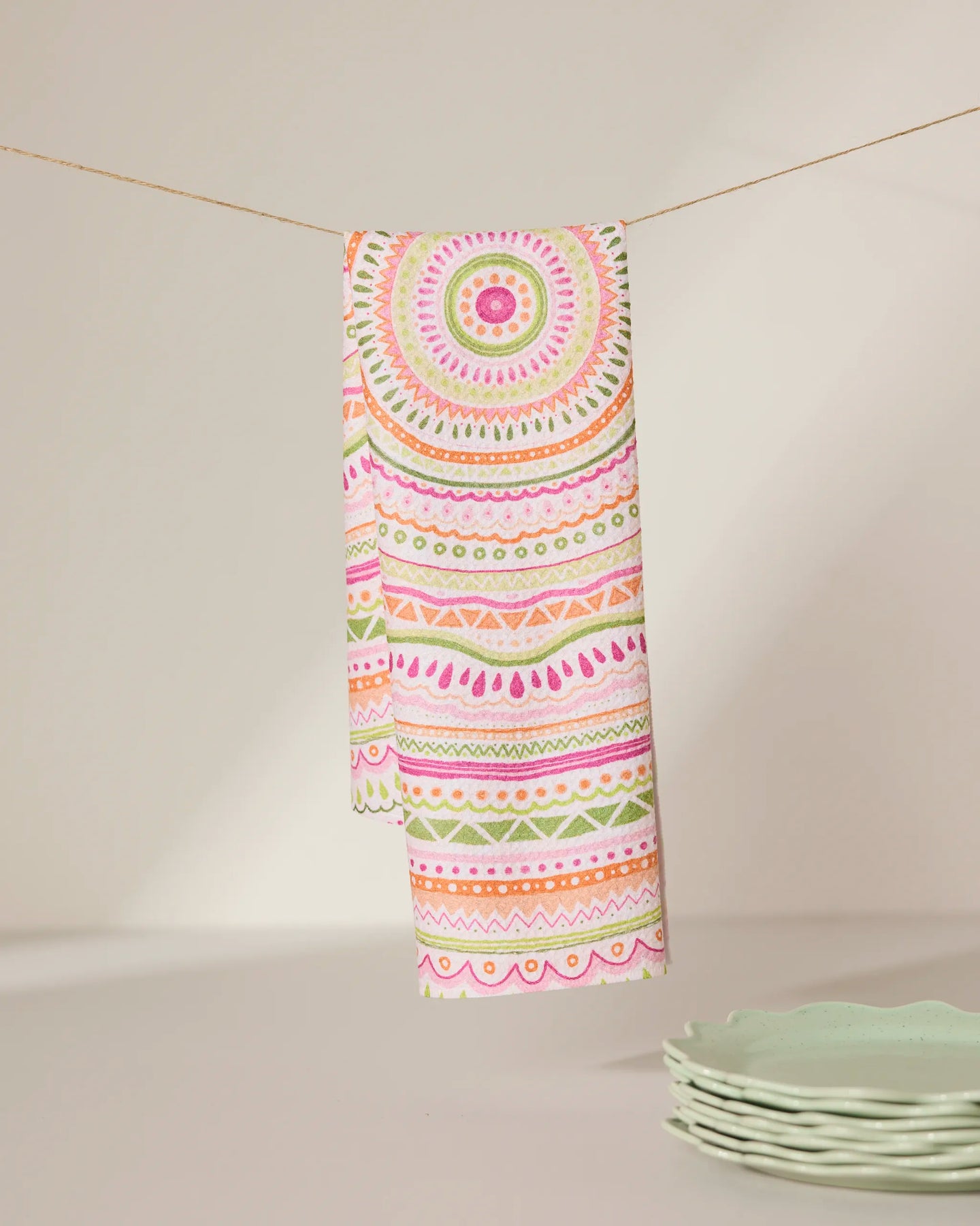 The GEOMETRY Bohemian Spring tea towel, crafted from recycled materials and adorned with vibrant pink, orange, and green mandala and geometric designs, hangs near a stack of light green plates on a white surface.