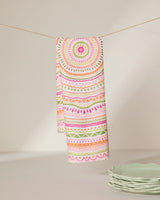 The GEOMETRY Bohemian Spring tea towel, crafted from recycled materials and adorned with vibrant pink, orange, and green mandala and geometric designs, hangs near a stack of light green plates on a white surface.