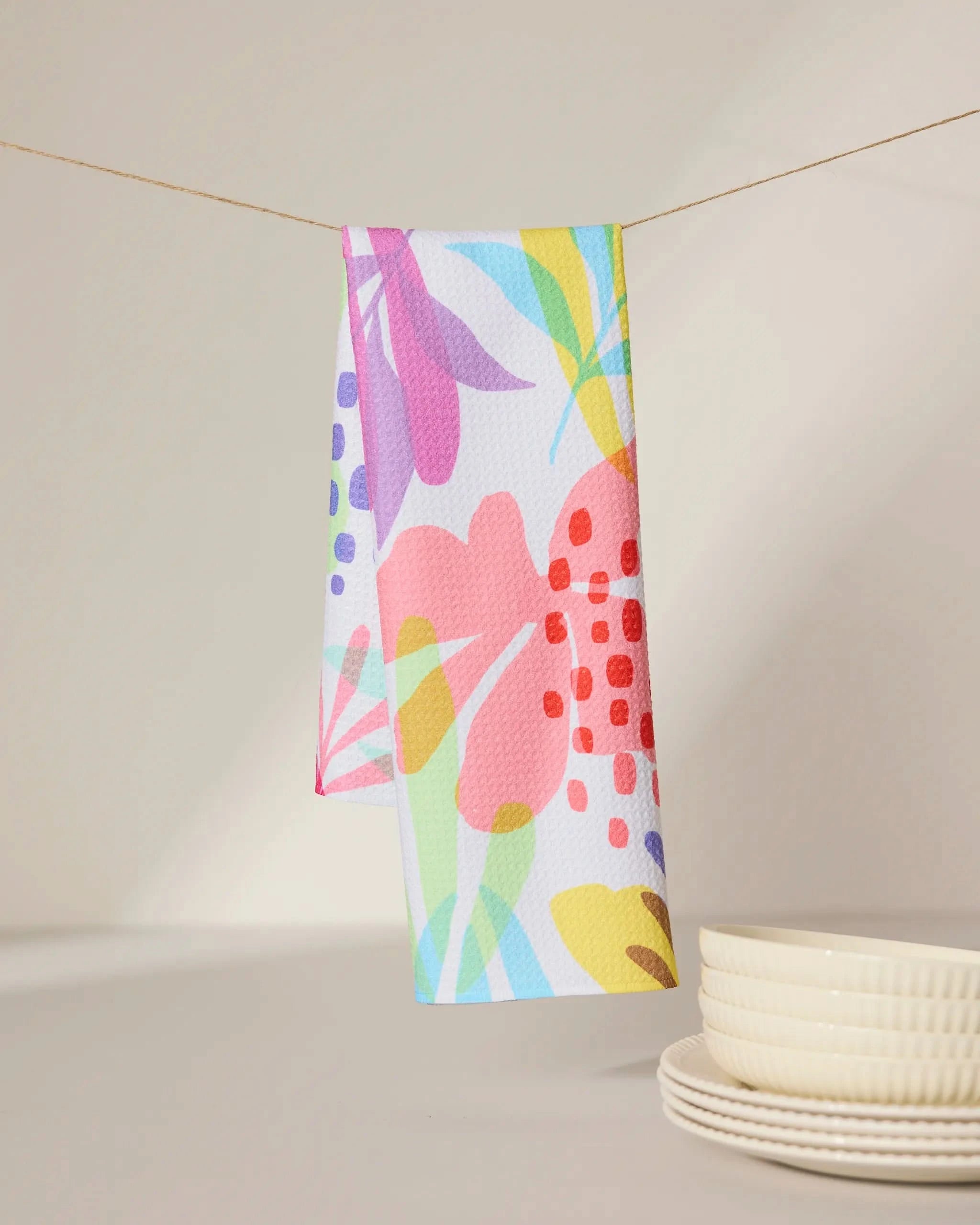 The GEOMETRY Botanical Burst tea towel, featuring vibrant abstract floral patterns, hangs on a string above a light surface beside a stack of textured white ceramic bowls.