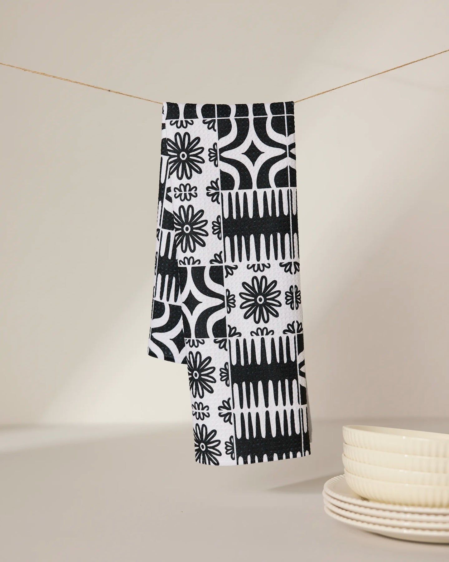 The GEOMETRY Bohemian Mosaic recycled kitchen towel, featuring a black and white pattern, hangs on a thin string above a beige surface, with a stack of white ceramic plates nearby.