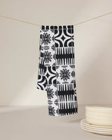 The GEOMETRY Bohemian Mosaic recycled kitchen towel, featuring a black and white pattern, hangs on a thin string above a beige surface, with a stack of white ceramic plates nearby.