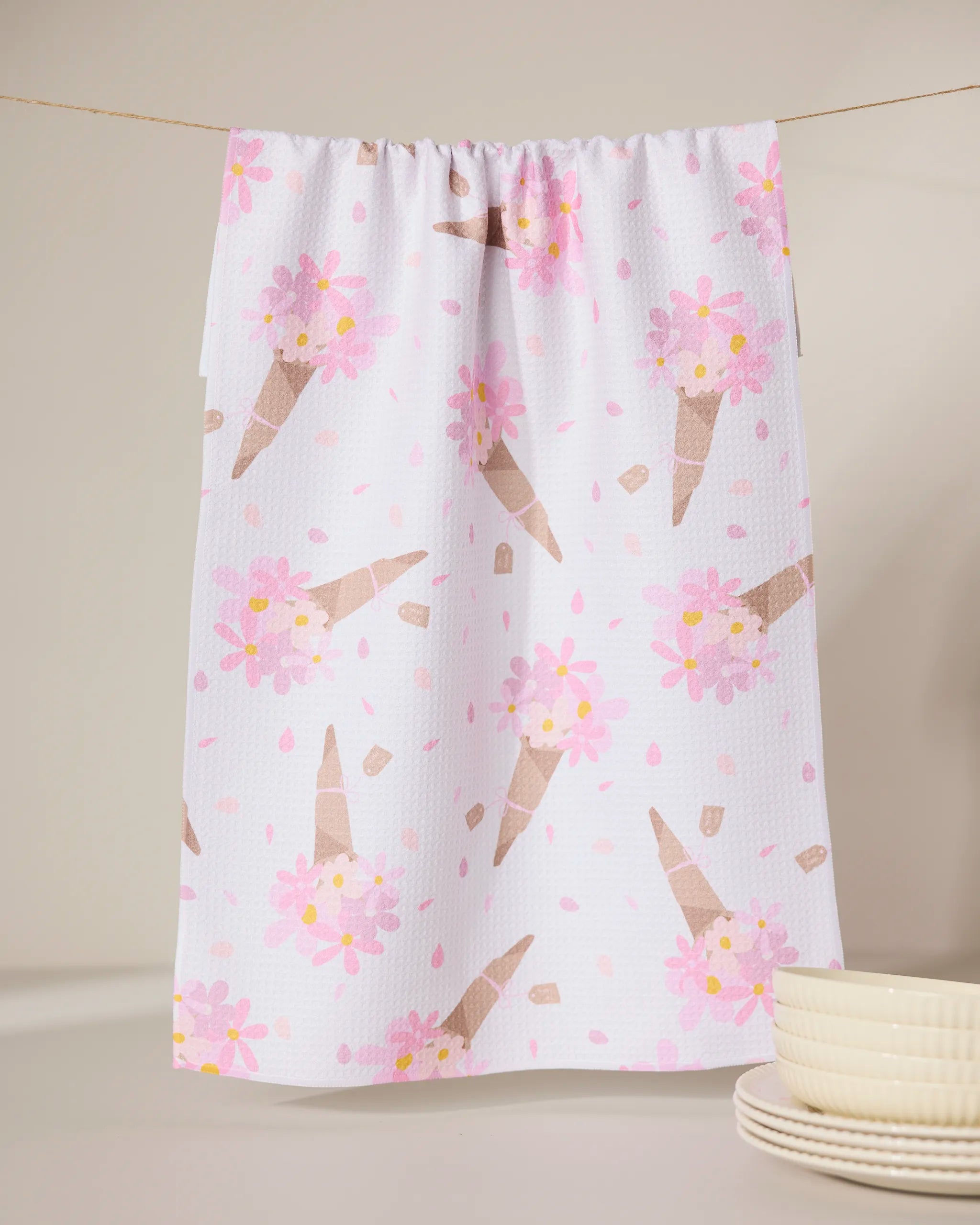 The GEOMETRY Gorgeous Gift is a white absorbent dish towel featuring a pastel pink and brown ice cream cone pattern, shown hanging on a clothesline with a stack of white plates in the corner.