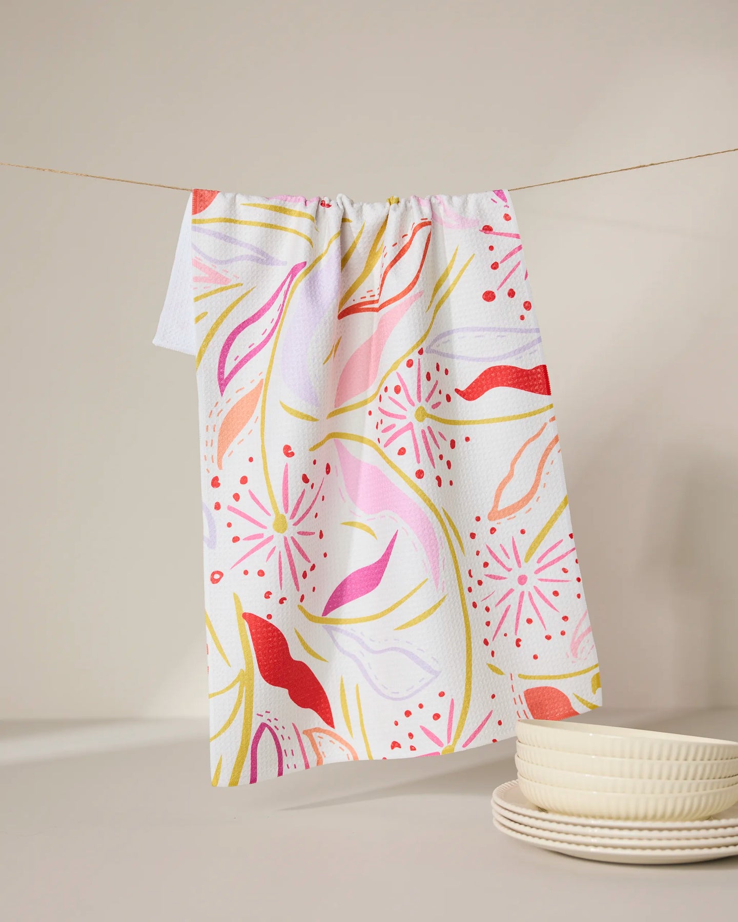 The GEOMETRY Grand Bloom kitchen towel, made from recycled materials, features vibrant pink, red, yellow, and orange patterns. Hanging on a line beside white textured plates on a softly lit surface, it brings color to any space.