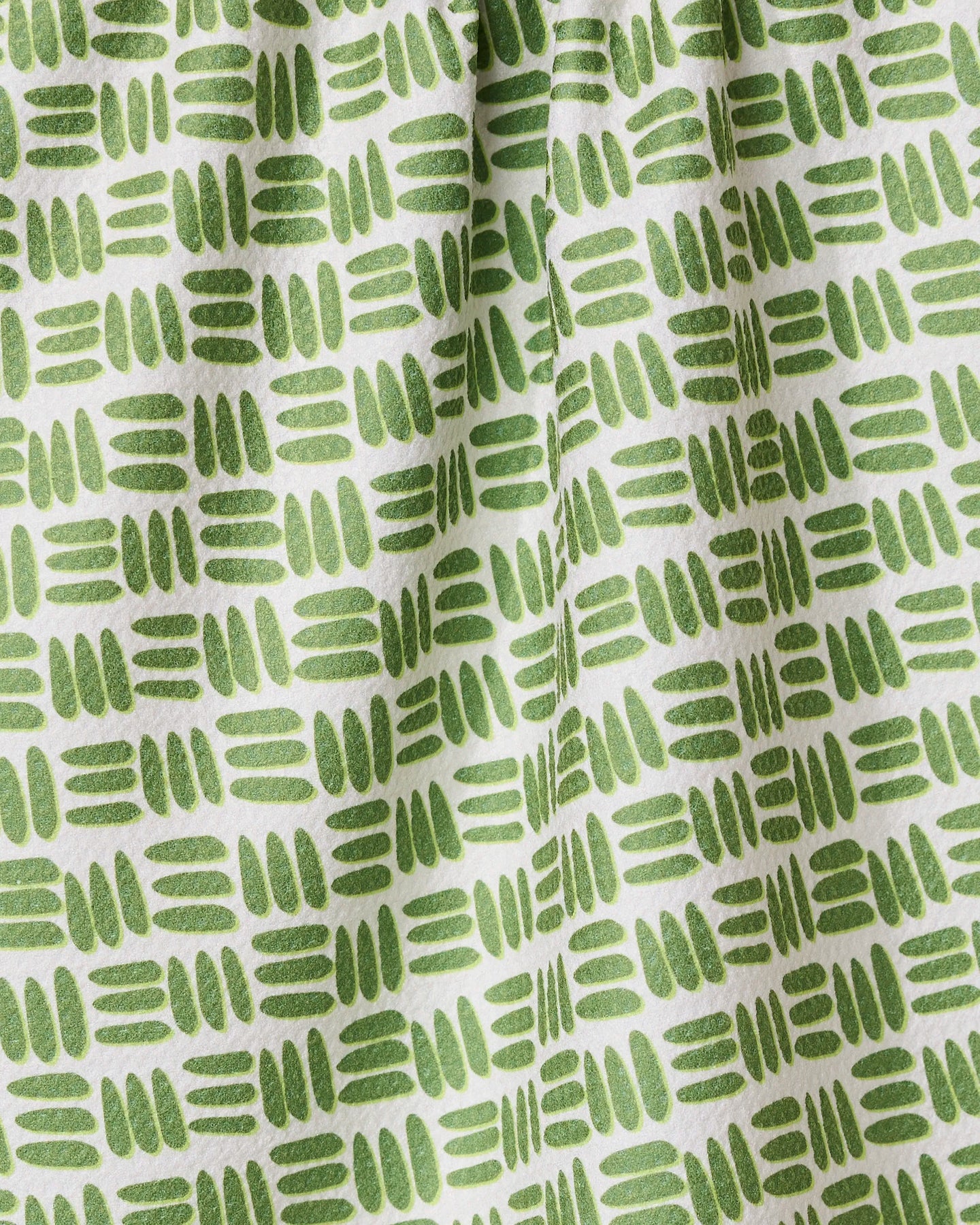 A close-up of the GEOMETRY Grass Hatch tea towel shows its ultra-absorbent white fabric with a repeating pattern of short green hand-drawn lines and ovals in horizontal rows, creating gentle folds and subtle shadows.