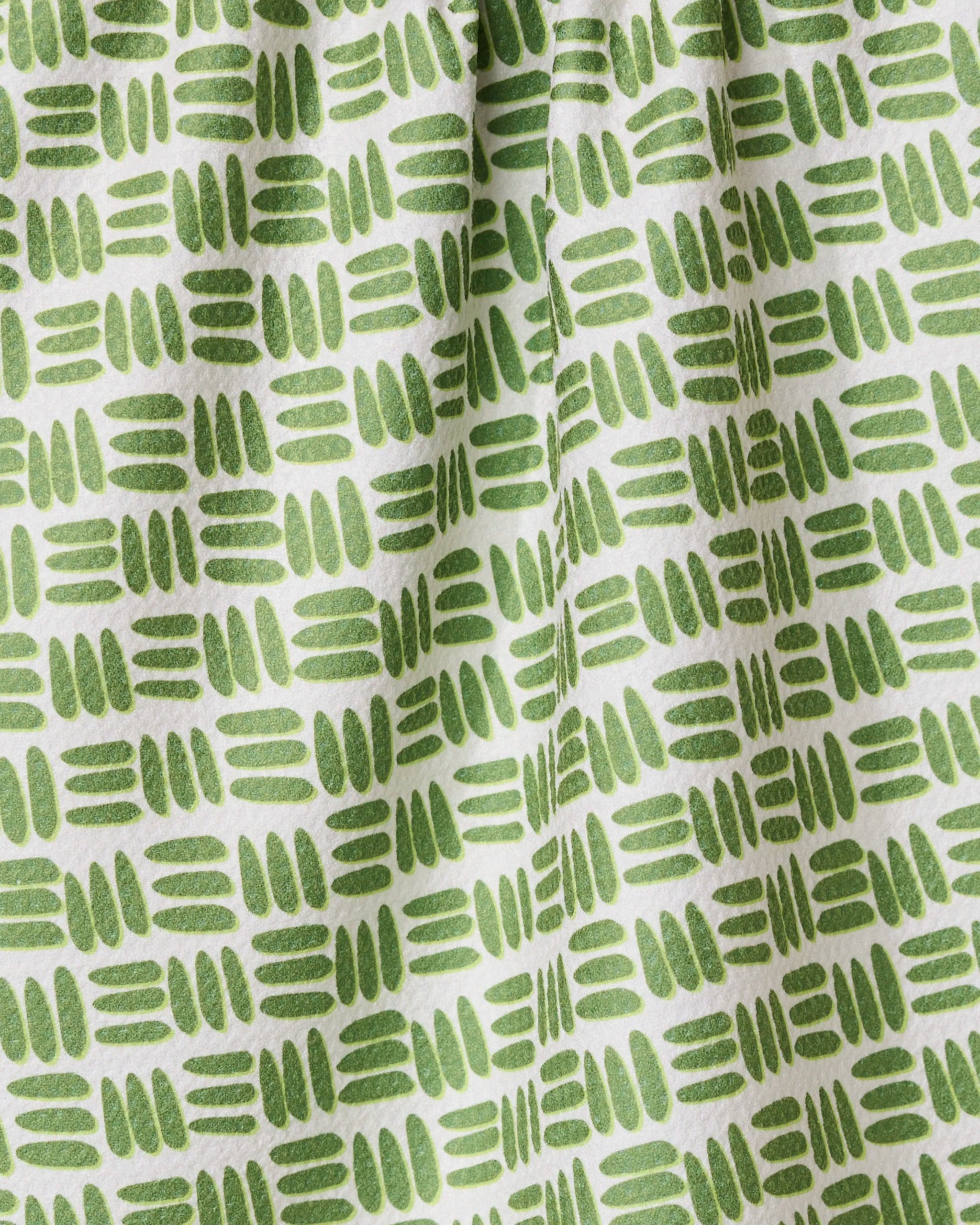 A close-up of the GEOMETRY Grass Hatch tea towel shows its ultra-absorbent white fabric with a repeating pattern of short green hand-drawn lines and ovals in horizontal rows, creating gentle folds and subtle shadows.