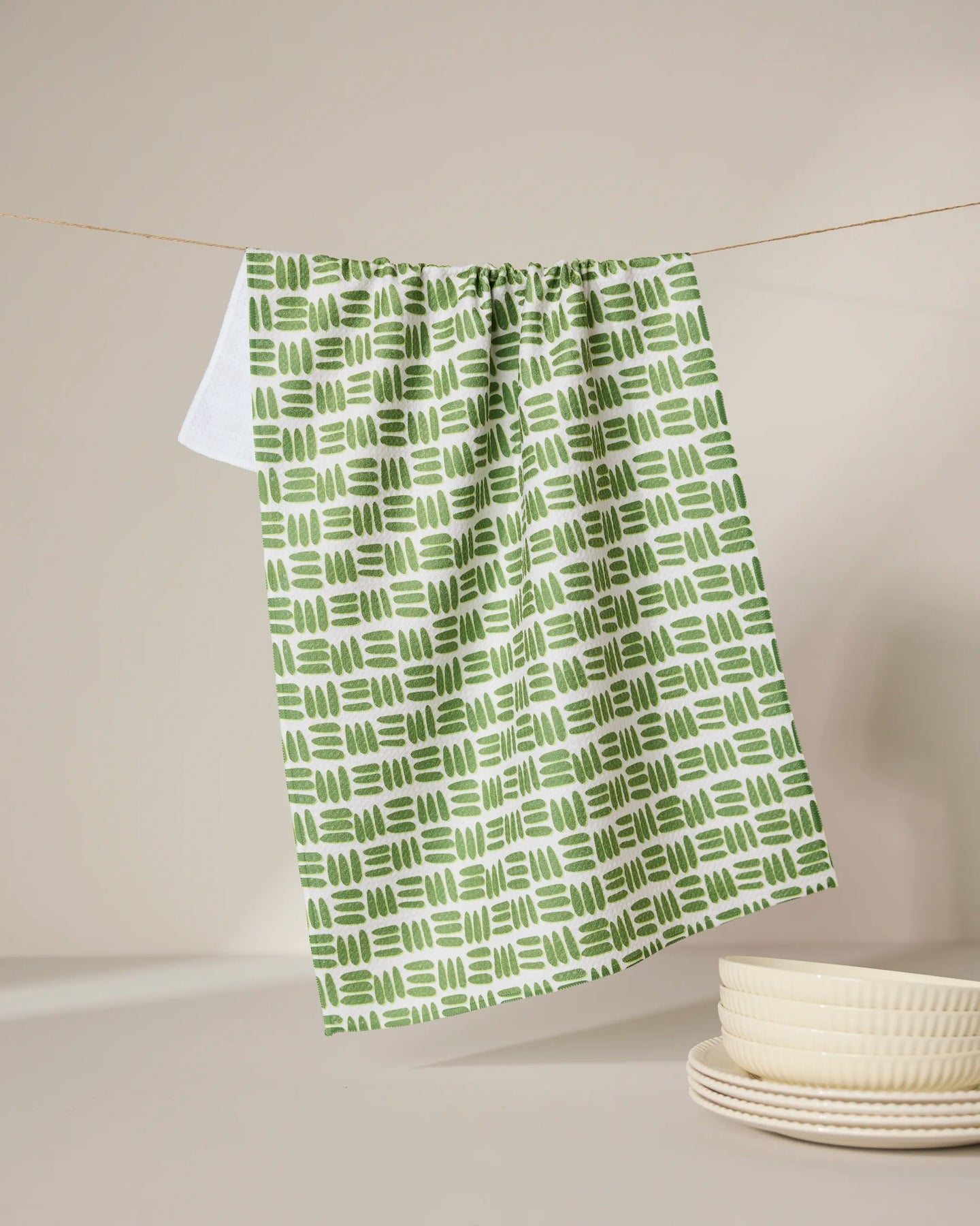 The GEOMETRY Grass Hatch tea towel, ultra-absorbent and featuring a green and white pattern, hangs on a clothesline above a white surface next to a stack of cream-colored dishes.