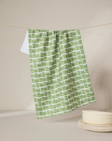 The GEOMETRY Grass Hatch tea towel, ultra-absorbent and featuring a green and white pattern, hangs on a clothesline above a white surface next to a stack of cream-colored dishes.