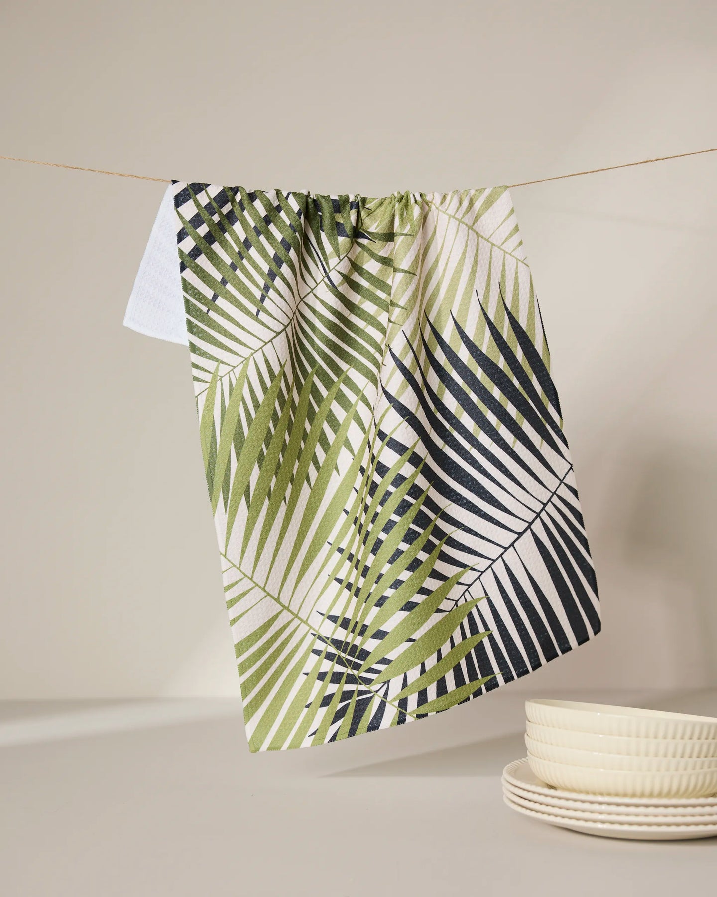 The GEOMETRY Green Fronds eco-friendly tea towel, featuring a green and black palm leaf design, hangs on a string above cream-colored plates against a neutral background.