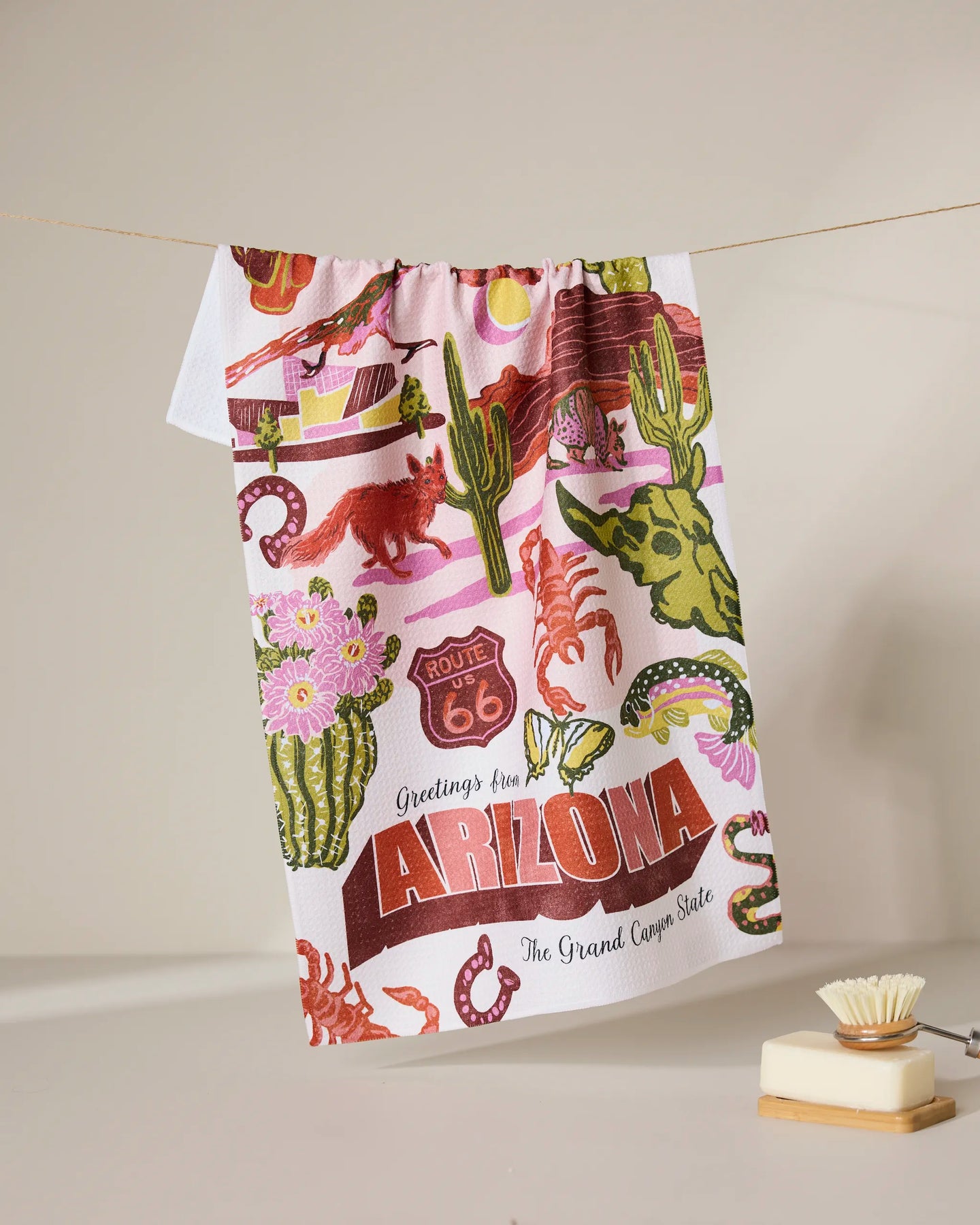 The Geometry Greetings From Arizona tea towel features cacti, Route 66 signs, desert wildlife, and a vibrant state slogan—perfect as a quick-drying kitchen towel or decorative accent.