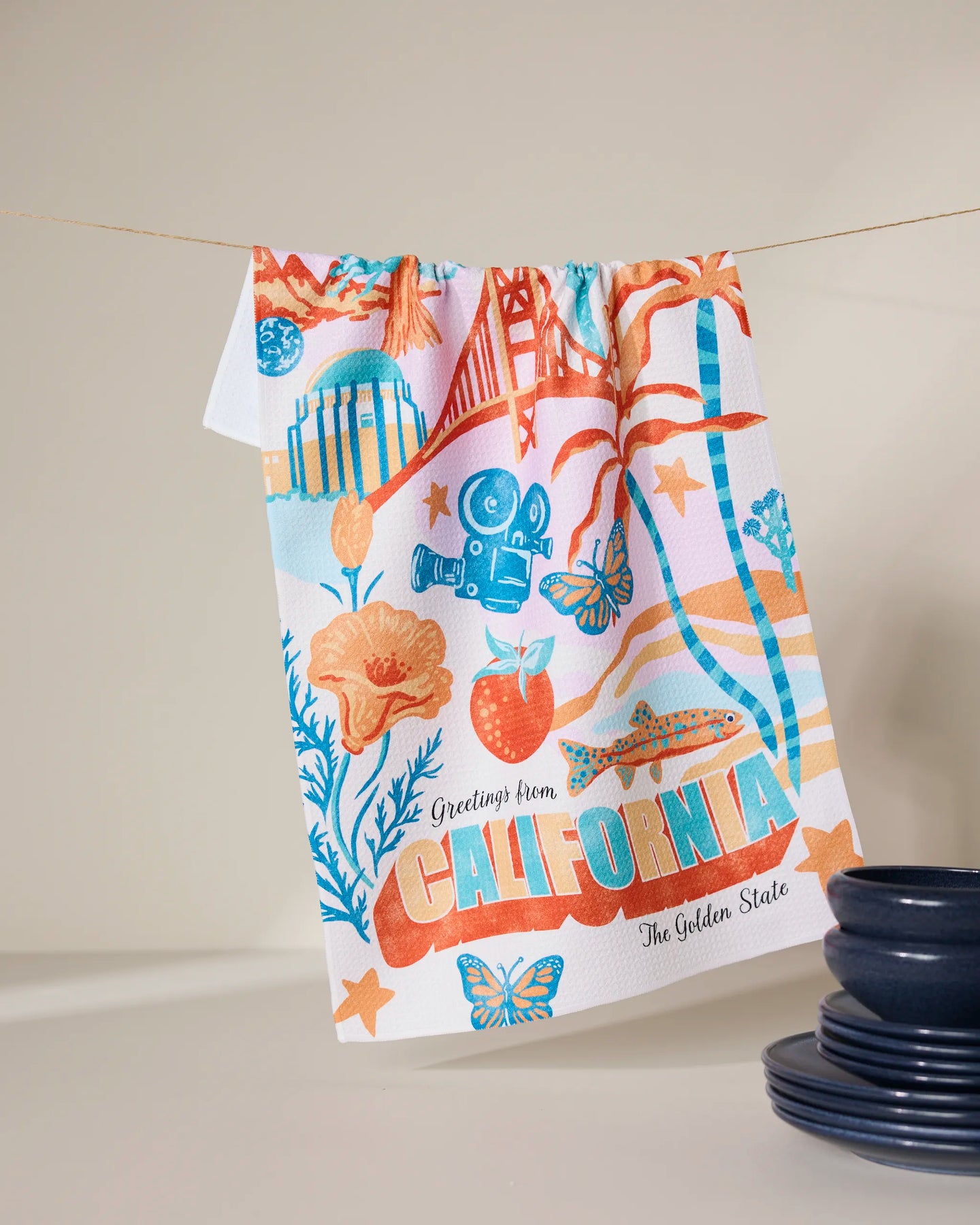 The Geometry Greetings From California tea towel is made from ultra-absorbent recycled materials and showcases colorful illustrations of California icons like the Golden Gate Bridge, poppies, and oranges.
