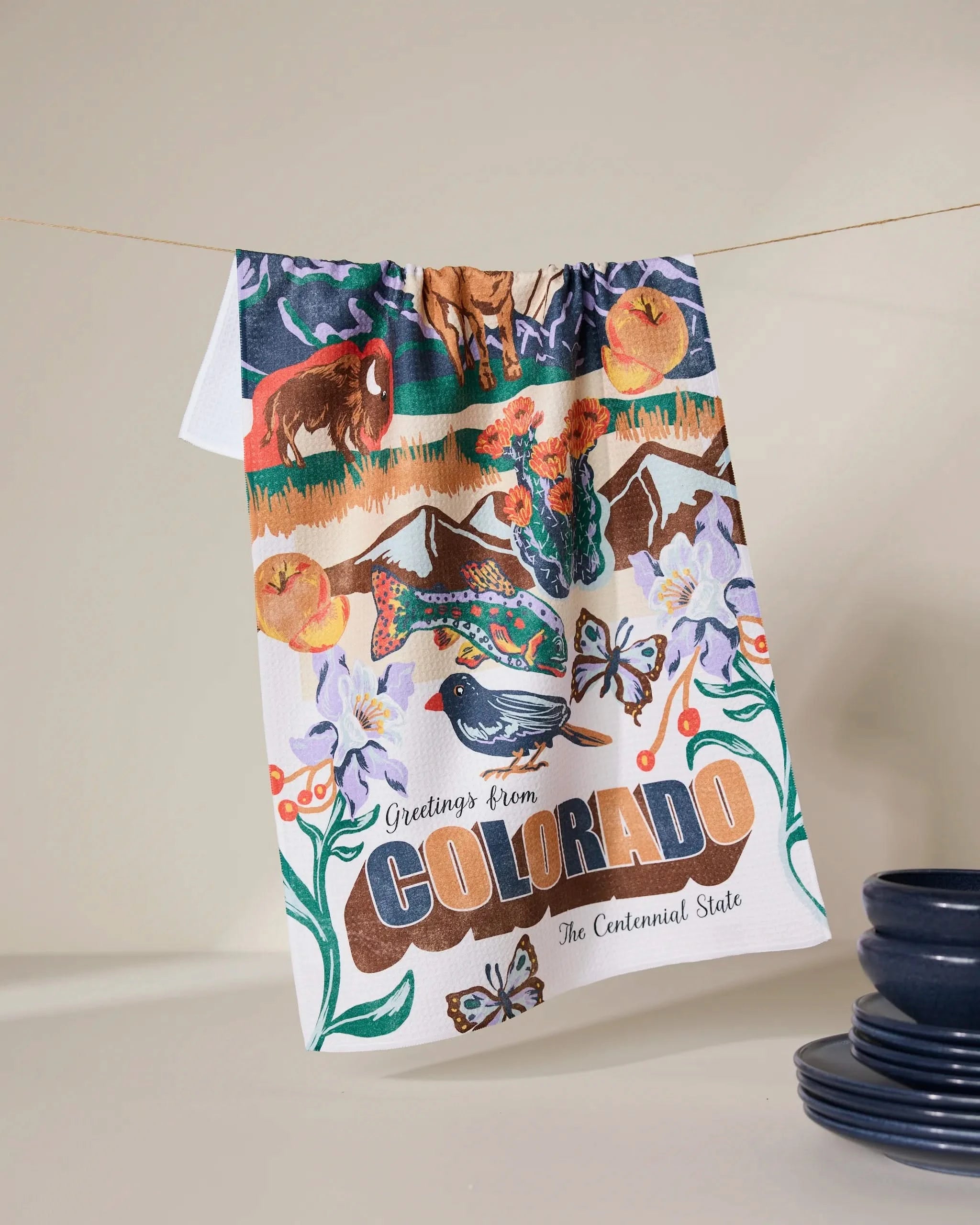Geometry’s “Greetings From Colorado” tea towel, quick-drying and colorful, hangs by blue dishes. It features Colorado wildlife, mountains, flowers, and “Greetings from Colorado, The Centennial State.”.