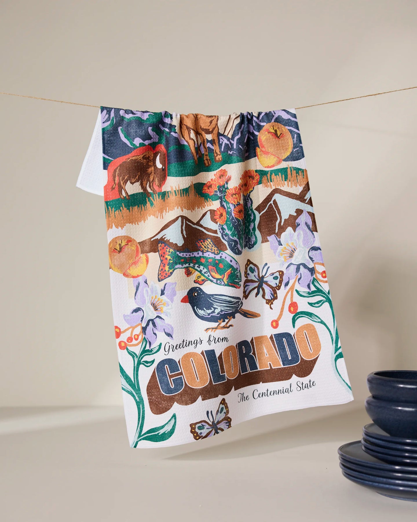 Geometry’s “Greetings From Colorado” tea towel, quick-drying and colorful, hangs by blue dishes. It features Colorado wildlife, mountains, flowers, and “Greetings from Colorado, The Centennial State.”.