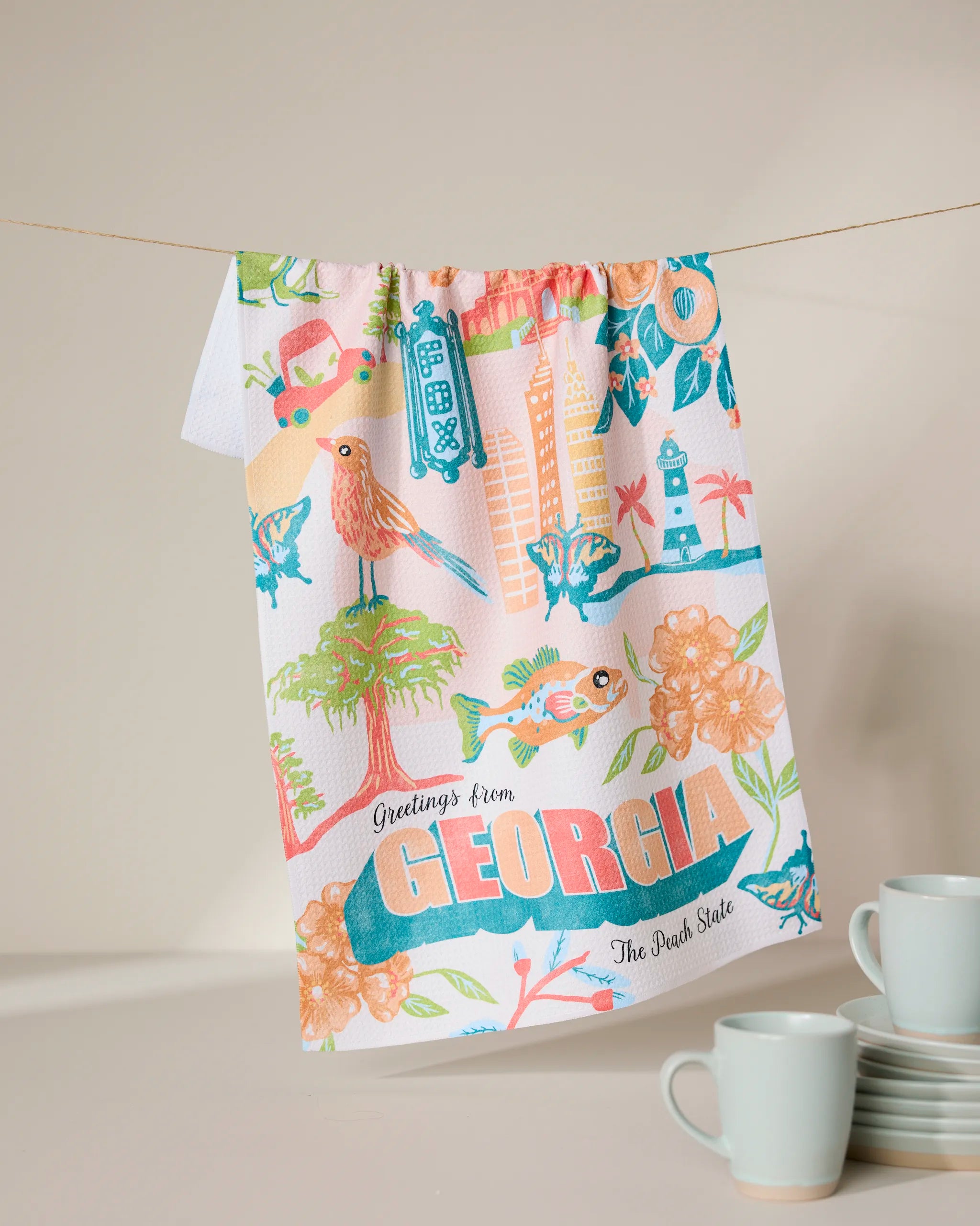 The Geometry Greetings From Georgia tea towel features colorful illustrations—birds, flowers, peaches, a fish, and landmarks—plus “Greetings from GEORGIA The Peach State.” Three matching mugs are stacked nearby.