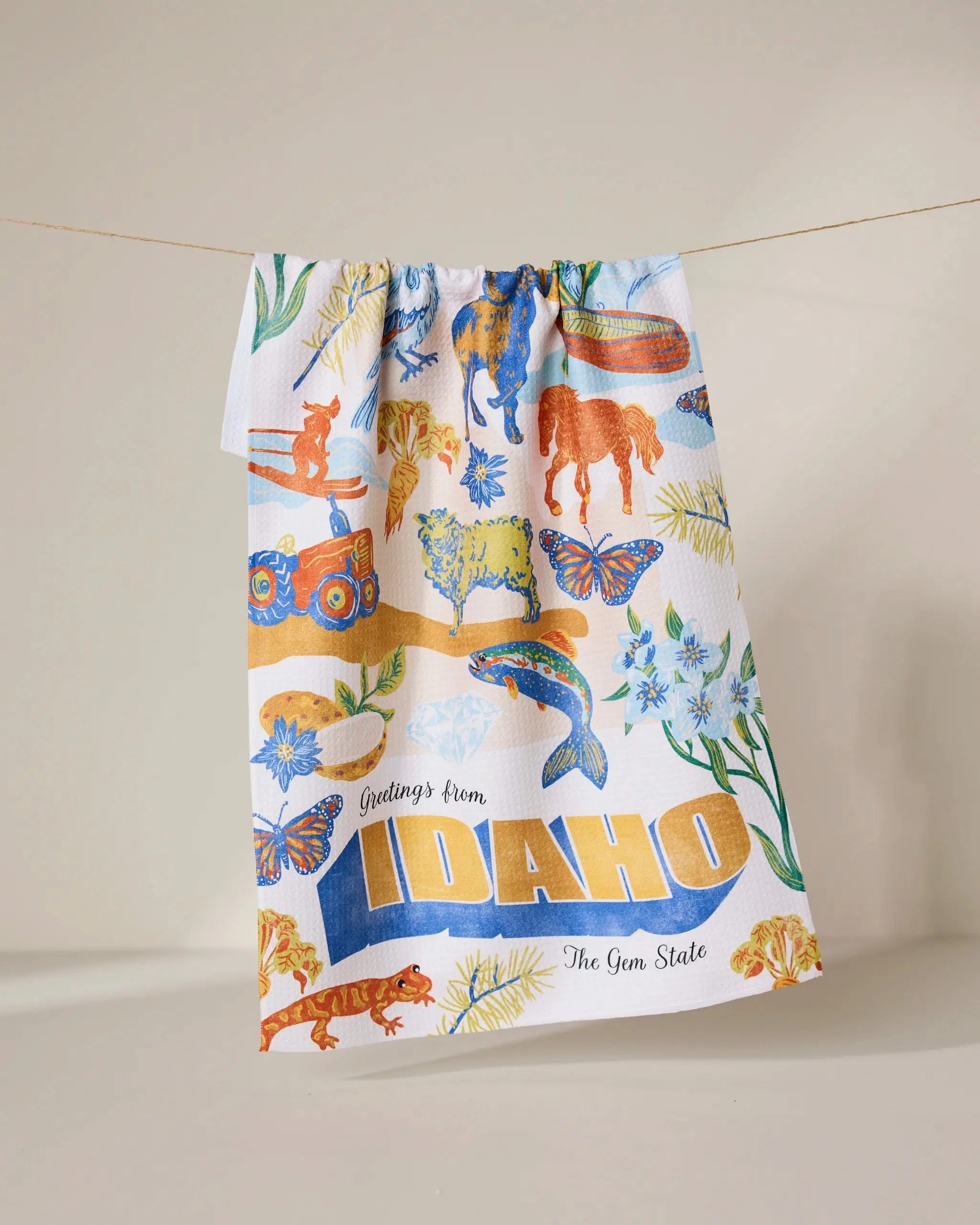 The Geometry Greetings From Idaho Kitchen Tea Towel hangs on a line, showcasing Idaho-themed wildlife, flowers, and scenery in colorful Geoweave fabric with bold yellow text: Greetings from IDAHO The Gem State.