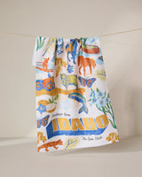 The Geometry Greetings From Idaho Kitchen Tea Towel hangs on a line, showcasing Idaho-themed wildlife, flowers, and scenery in colorful Geoweave fabric with bold yellow text: Greetings from IDAHO The Gem State.