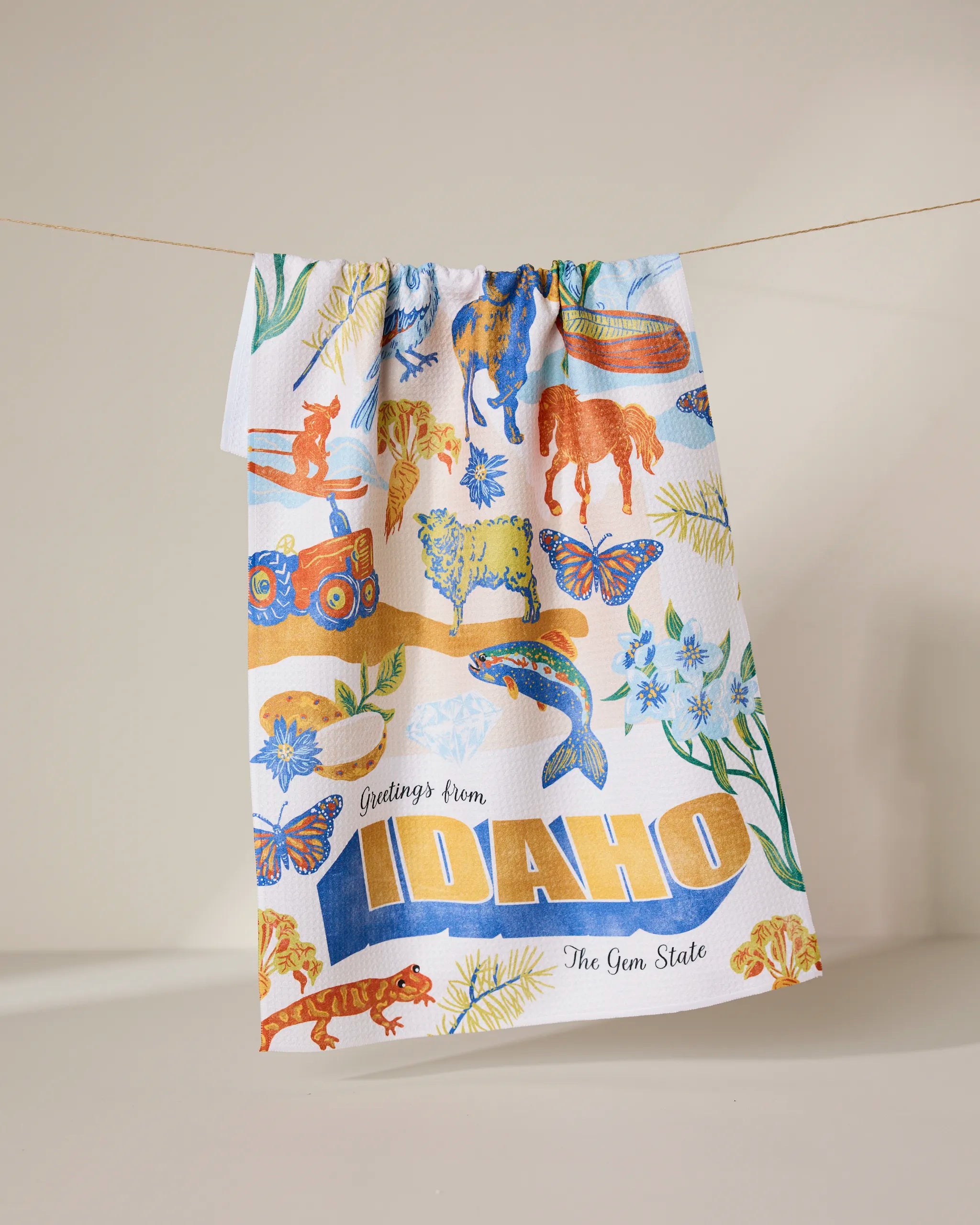 The Geometry Greetings From Idaho Kitchen Tea Towel hangs on a line, showcasing Idaho-themed wildlife, flowers, and scenery in colorful Geoweave fabric with bold yellow text: Greetings from IDAHO The Gem State.
