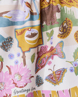 The Geometry Greetings From Montana tea towel features a colorful design with Montana-themed elements like a bear, yellow welcome sign, fish, pinecones, wildflowers, butterfly, and Greetings from Montana text—printed on quick-drying fabric.
