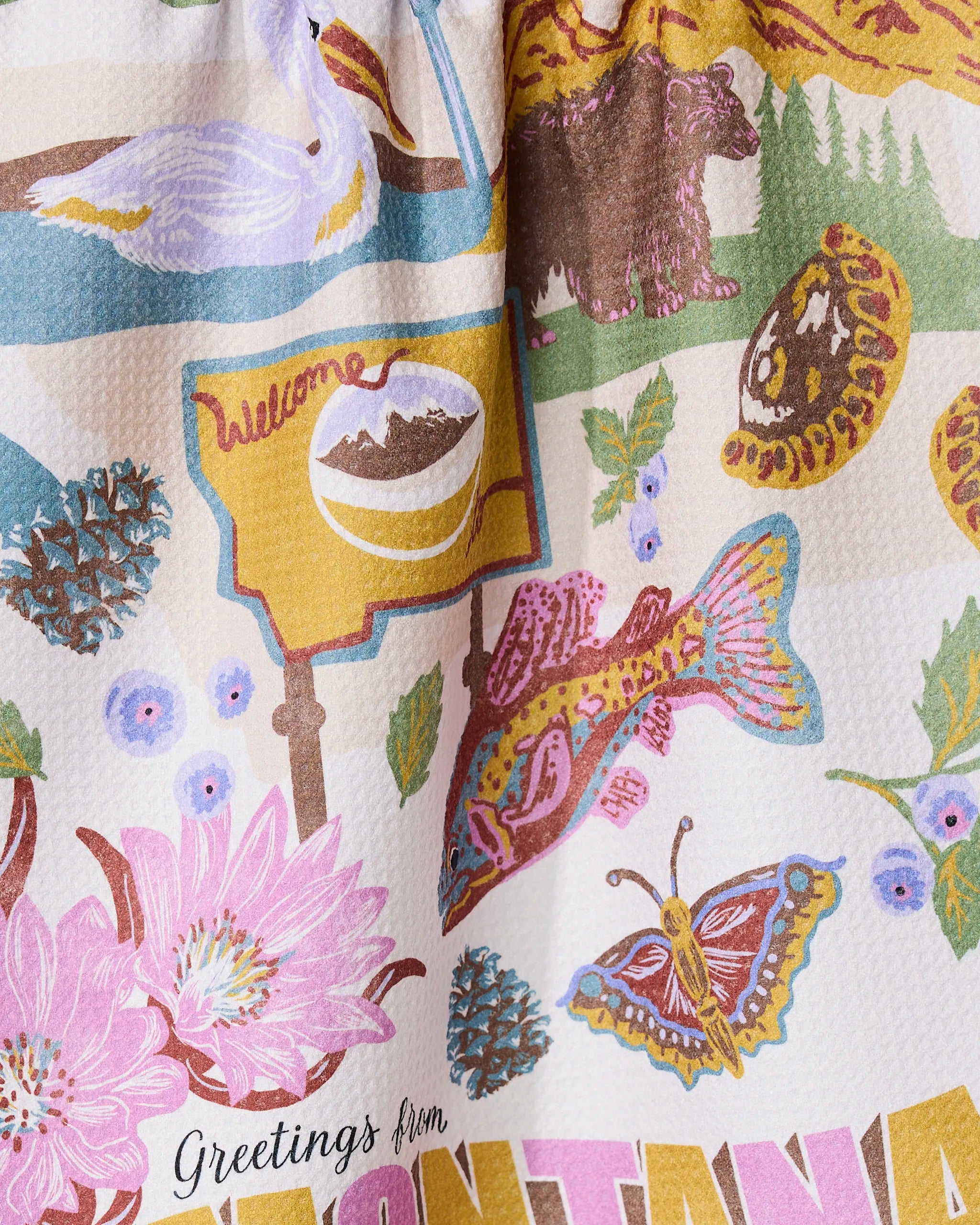 The Geometry Greetings From Montana tea towel features a colorful design with Montana-themed elements like a bear, yellow welcome sign, fish, pinecones, wildflowers, butterfly, and Greetings from Montana text—printed on quick-drying fabric.