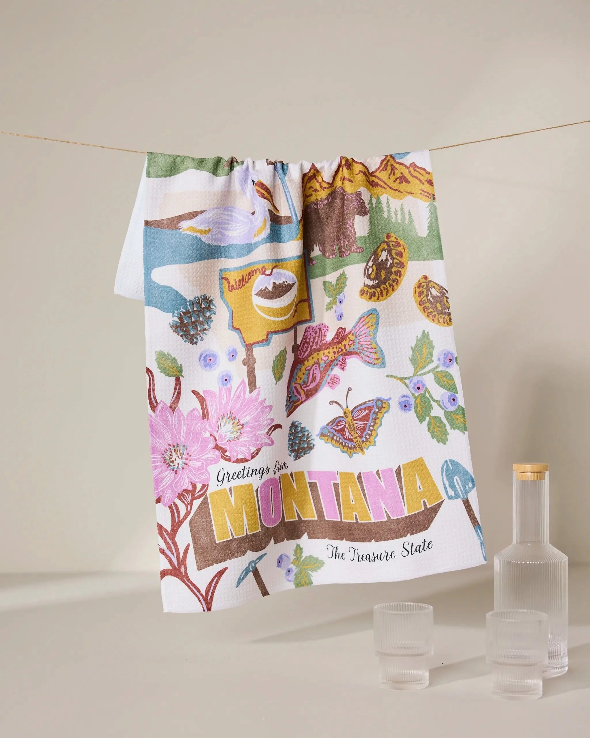 The Geometry Greetings From Montana tea towel, made from recycled materials, displays illustrated landmarks—flowers, trout, mountains, pine cones—with Greetings from Montana The Treasure State. Three glass containers accompany it.