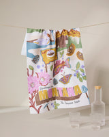 The Geometry Greetings From Montana tea towel, made from recycled materials, displays illustrated landmarks—flowers, trout, mountains, pine cones—with Greetings from Montana The Treasure State. Three glass containers accompany it.