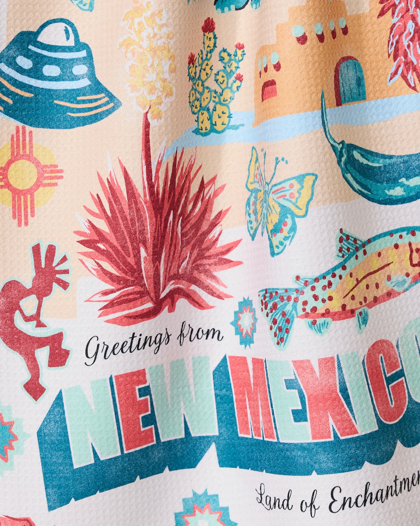 Geometry’s “Greetings From New Mexico” dish towel features a vibrant design with New Mexico icons—UFO, cactus, chili pepper, Kokopelli, fish, adobe building, and Zia sun symbol—plus the text “Greetings from NEW MEXICO Land of Enchantment.”.