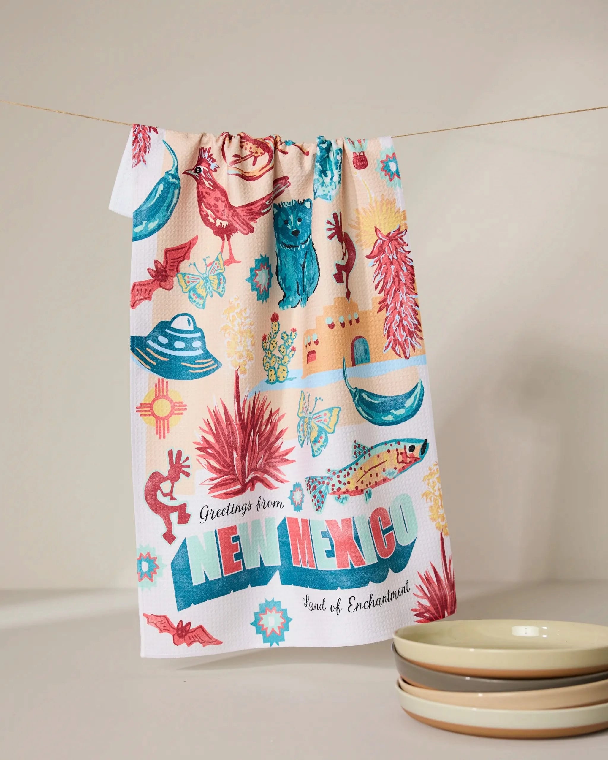 The Geometry Greetings From New Mexico dish towel hangs on a line, decorated with roadrunners, cacti, chili peppers, and the phrase “Greetings from New Mexico: Land of Enchantment.” Three stacked bowls are nearby.