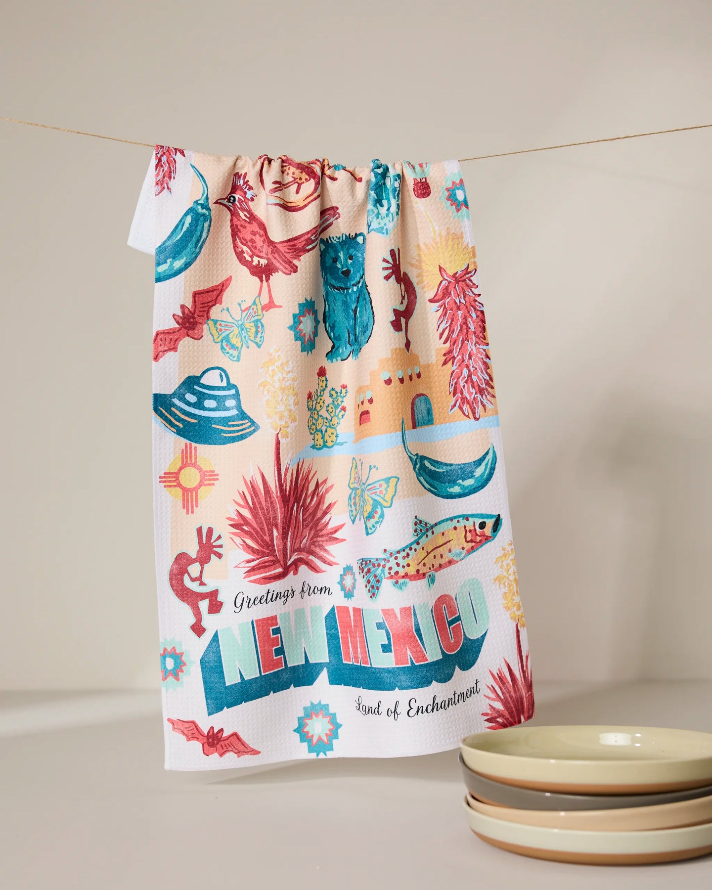 The Geometry Greetings From New Mexico dish towel hangs on a line, decorated with roadrunners, cacti, chili peppers, and the phrase “Greetings from New Mexico: Land of Enchantment.” Three stacked bowls are nearby.