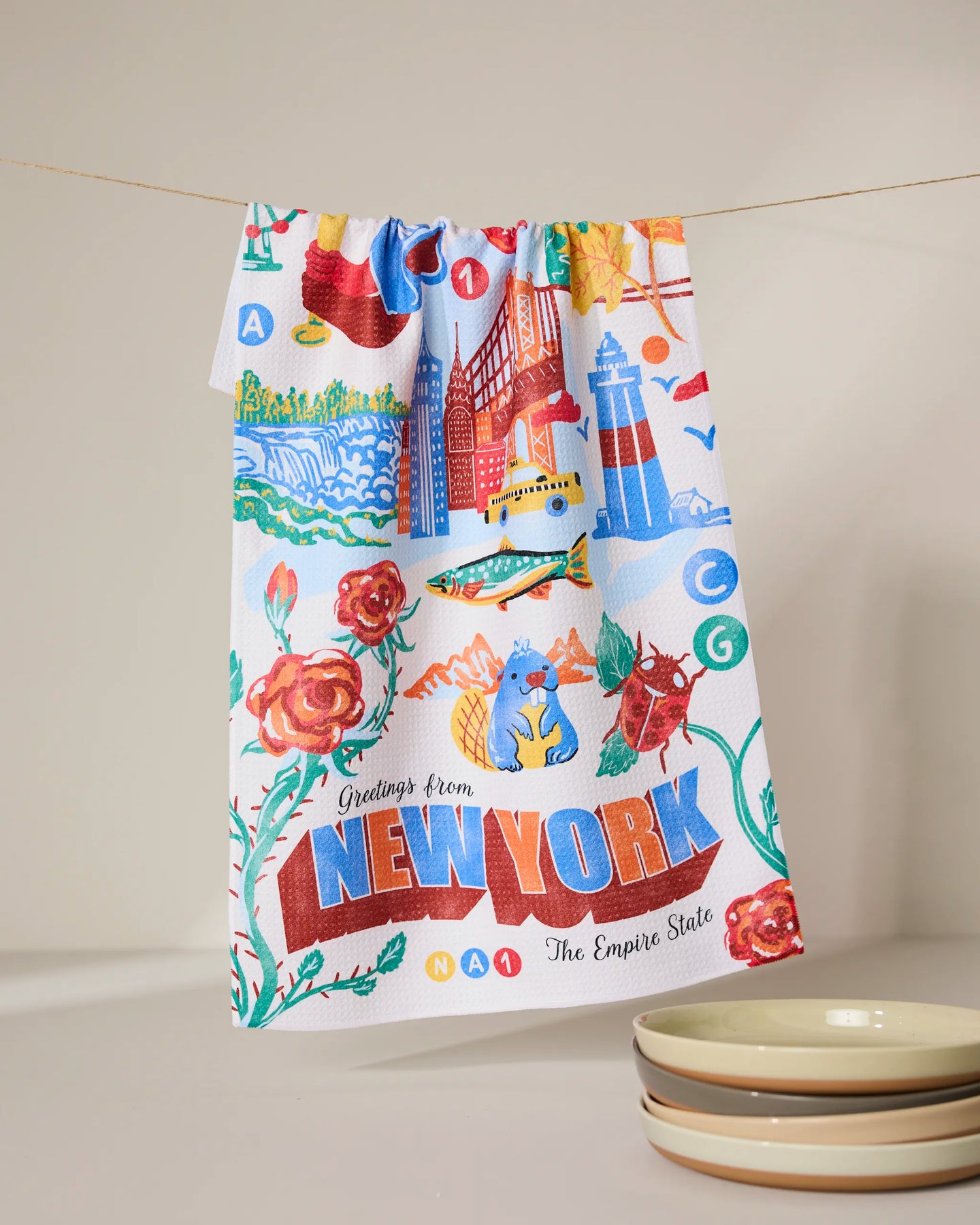 The Geometry Greetings From New York kitchen towel, made from recycled materials and featuring New York landmarks, roses, a blue cat, and text, hangs on a line beside three stacked plates.
