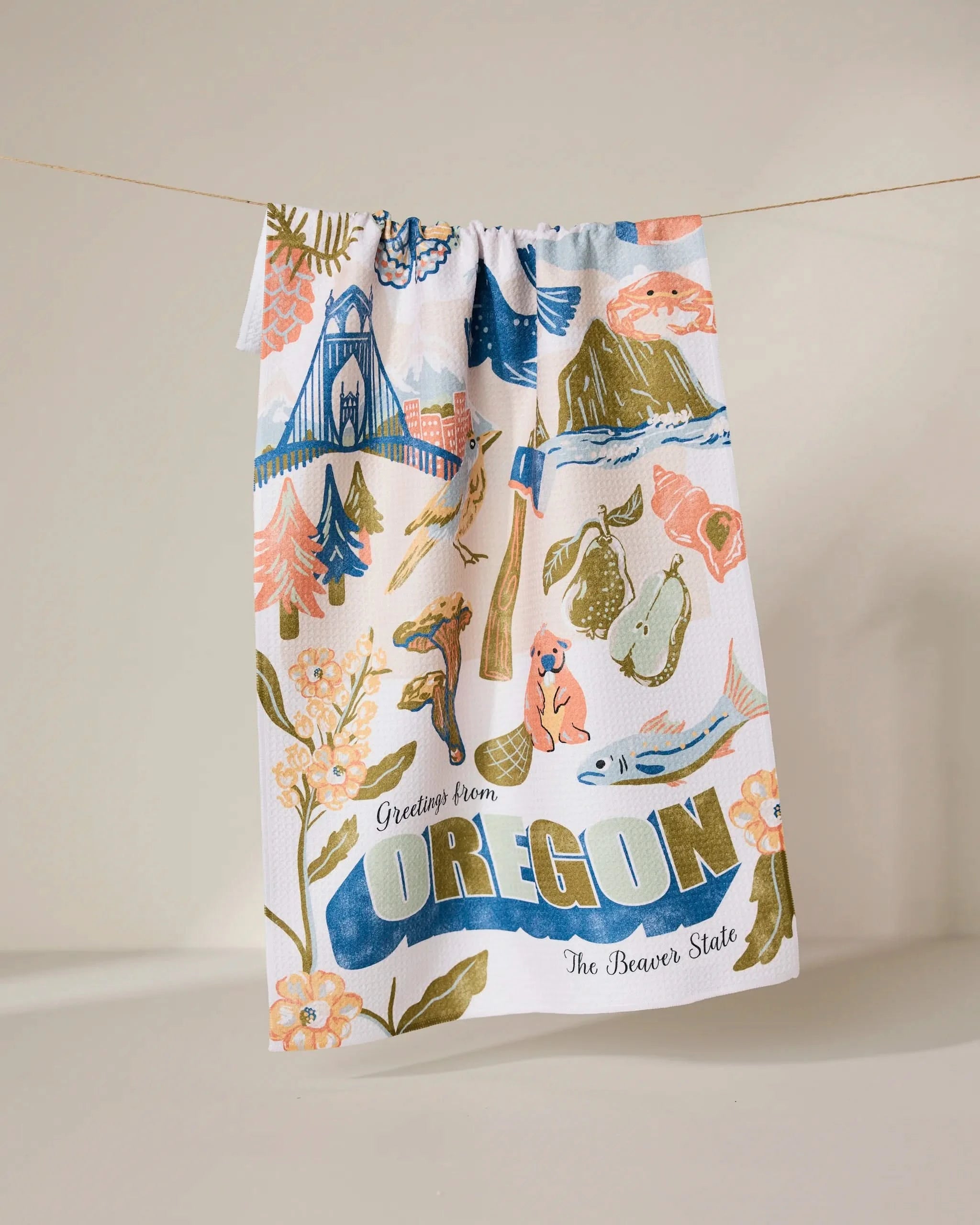 The Geometry Greetings From Oregon recycled kitchen towel hangs on a line, decorated with colorful Oregon landmarks, nature, animals, and bold “Greetings from OREGON The Beaver State” text among flowers, trees, and scenic icons.