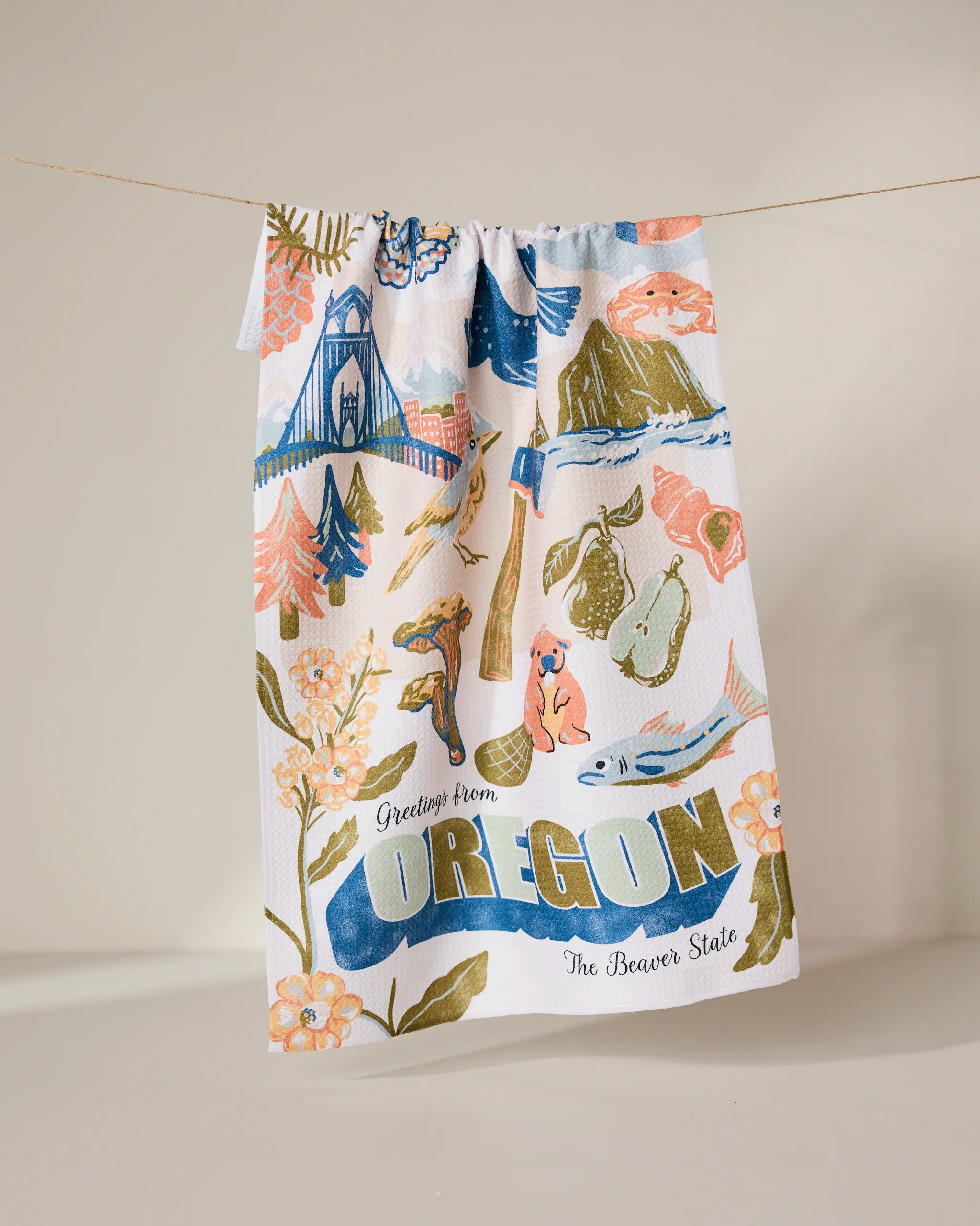 The Geometry Greetings From Oregon recycled kitchen towel hangs on a line, decorated with colorful Oregon landmarks, nature, animals, and bold “Greetings from OREGON The Beaver State” text among flowers, trees, and scenic icons.