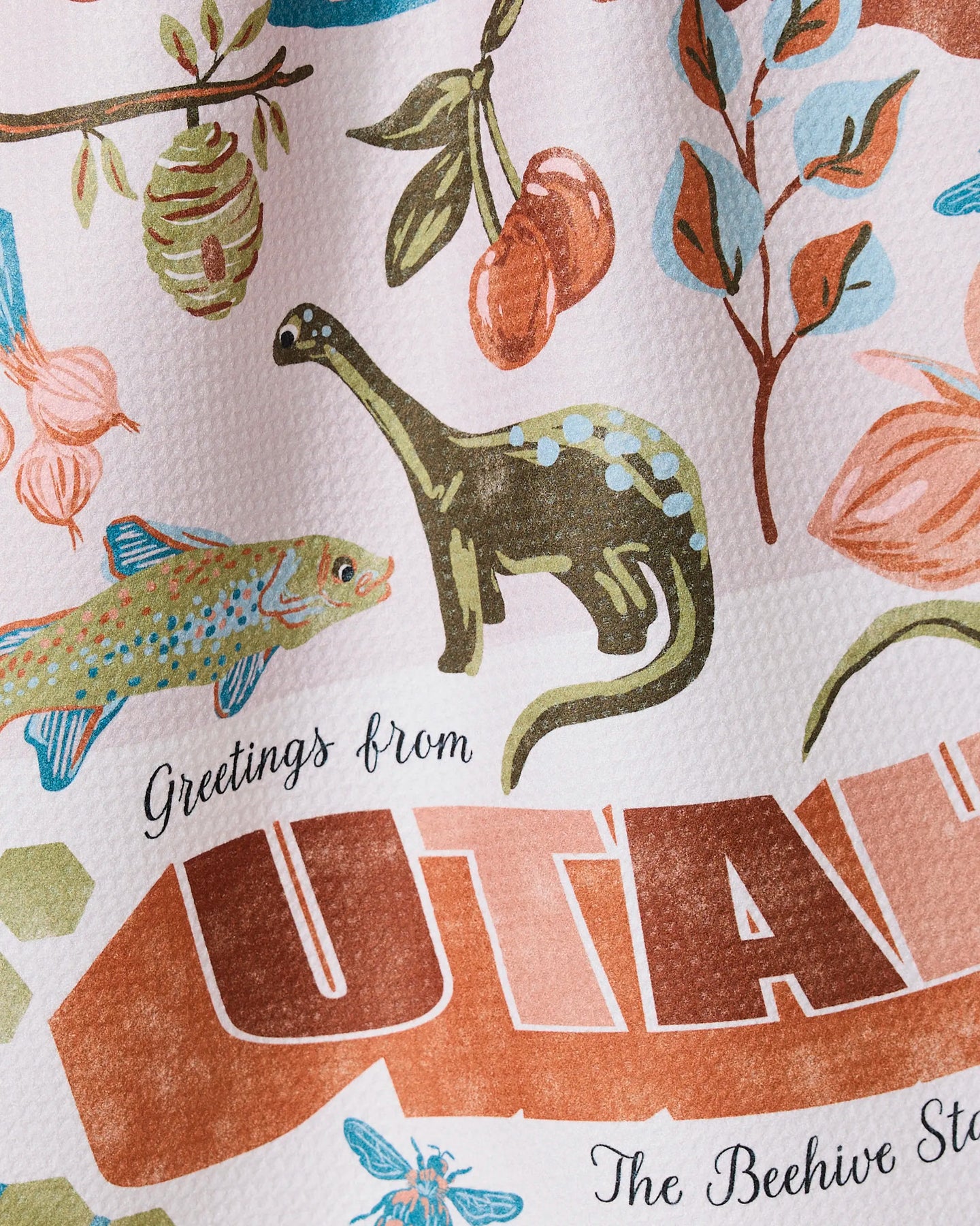 A close-up of the Greetings From Utah tea towel by Geometry, featuring a vibrant illustrated design with a dinosaur, fish, cherries, beehive, and leaves on textured ultra-absorbent fabric.